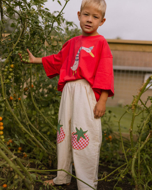 A young boy stands in a tomato garden wearing a red oversized t-shirt with a cartoon bird graphic and cream-colored linen pants featuring two strawberry appliques on the knees.