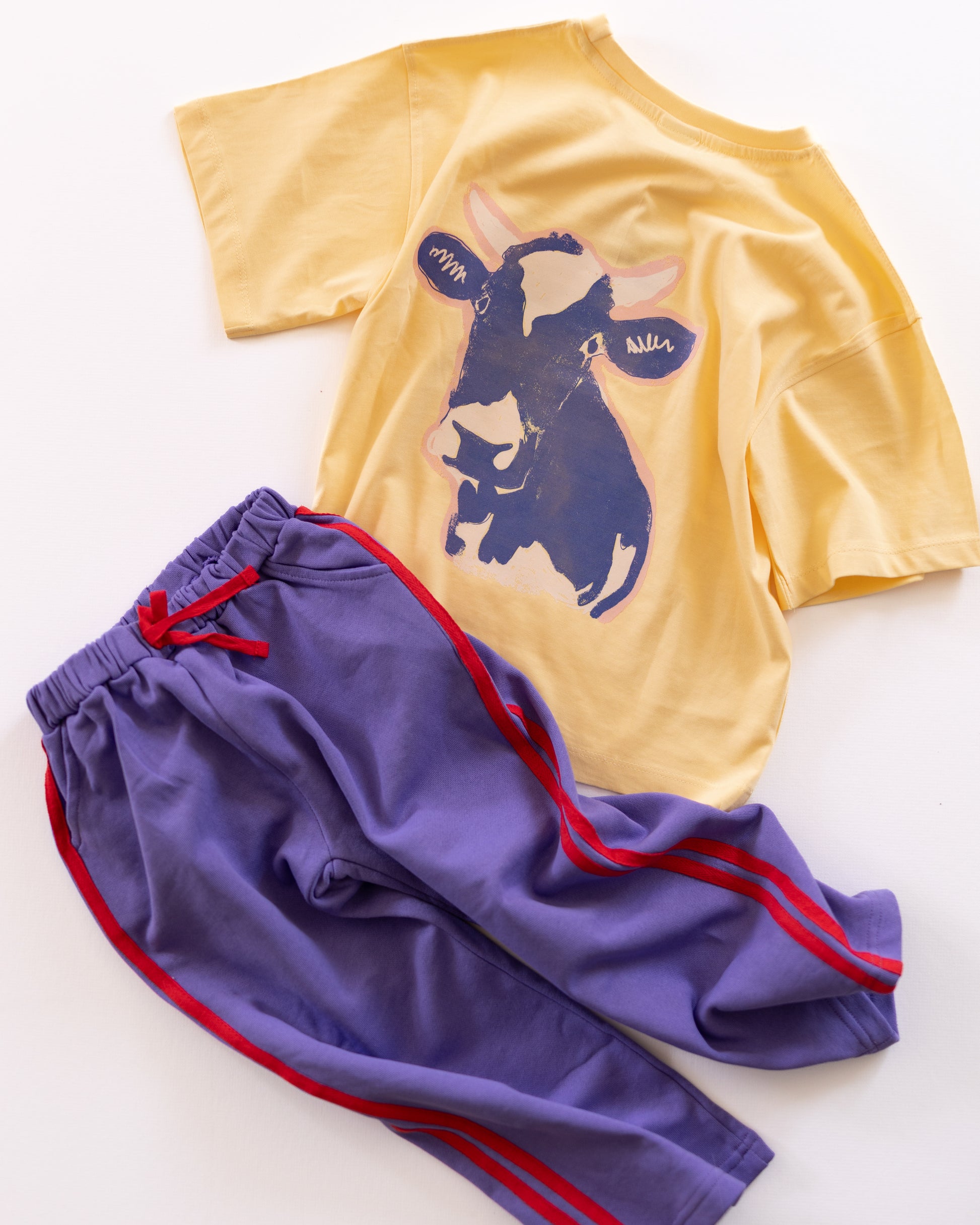 A children's two-piece outfit laid flat on a white background, featuring a yellow short-sleeve T-shirt with a large cow graphic and purple drawstring pants with red side stripes.