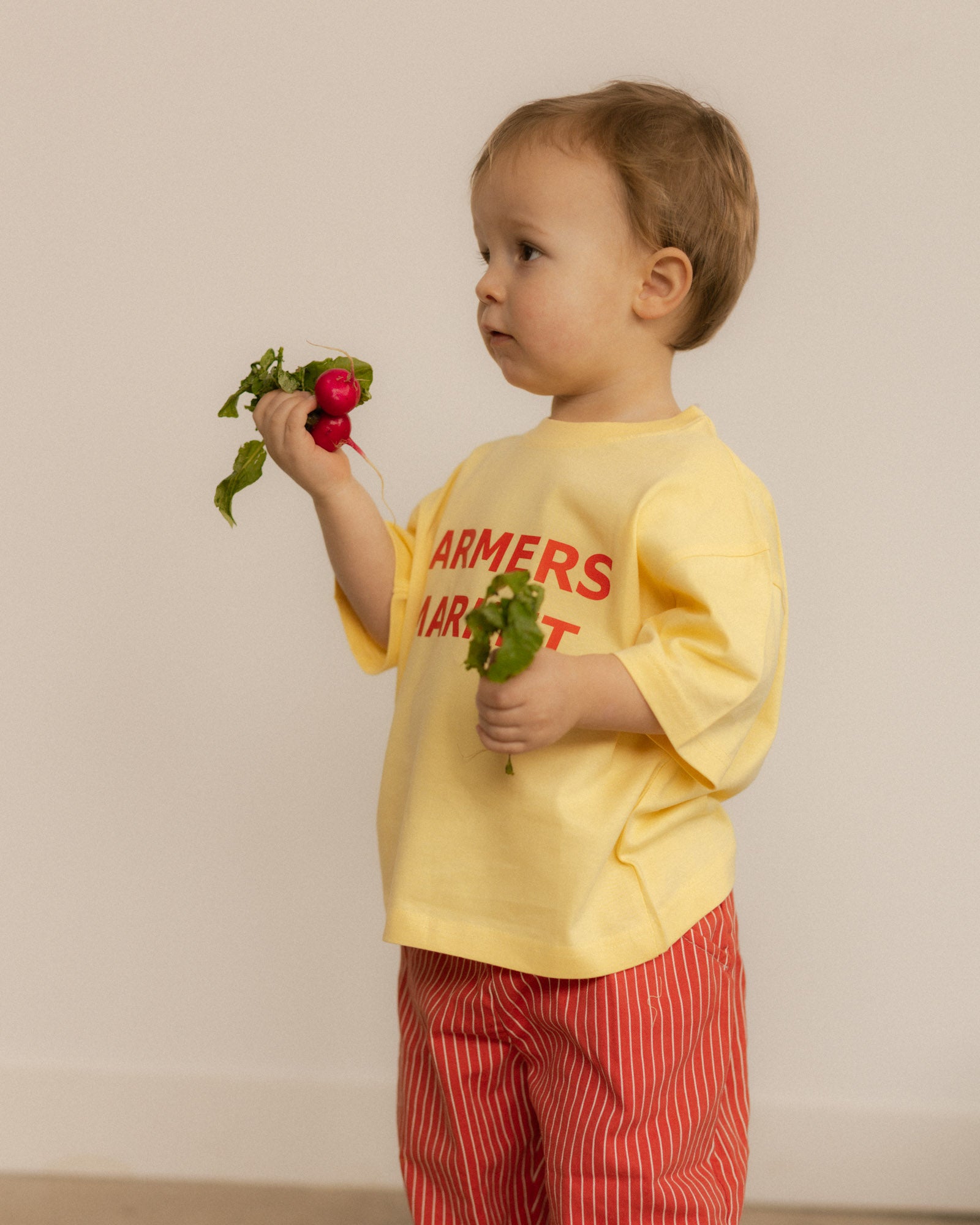 Child wearing a yellow shirt with 'Farmers Market' text, holding vegetables against a plain background