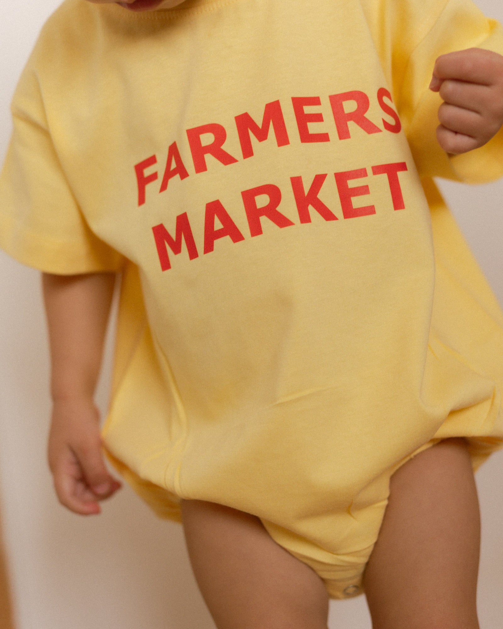 Yellow romper with 'Farmers Market' text worn by a child against a plain background