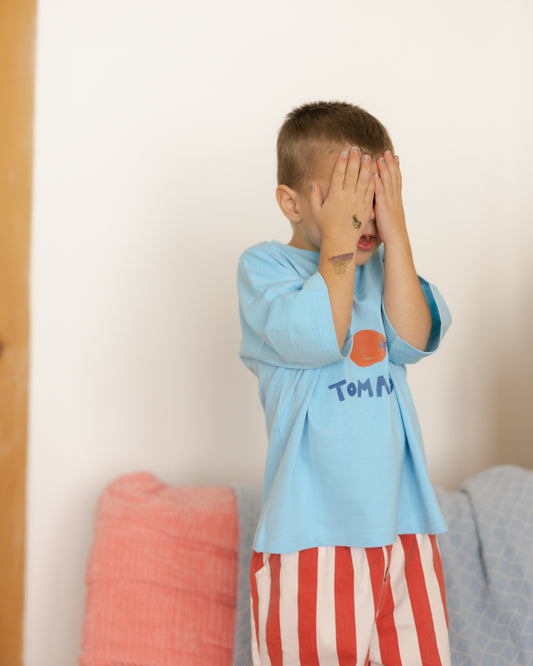 child wearing a blue shirt with red tomatoes graphic on it. hiding behind his hands.
