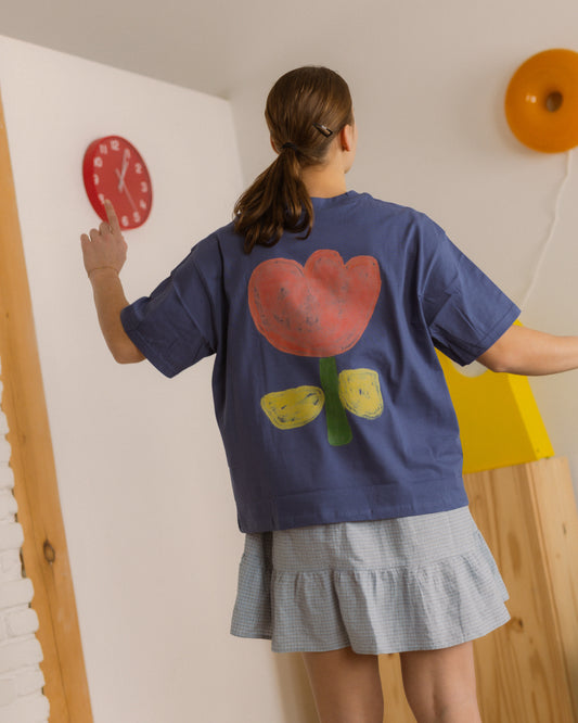 Person wearing a blue sweatshirt with a colorful flower design on the back, standing in a room with a clock and orange object on the wall.