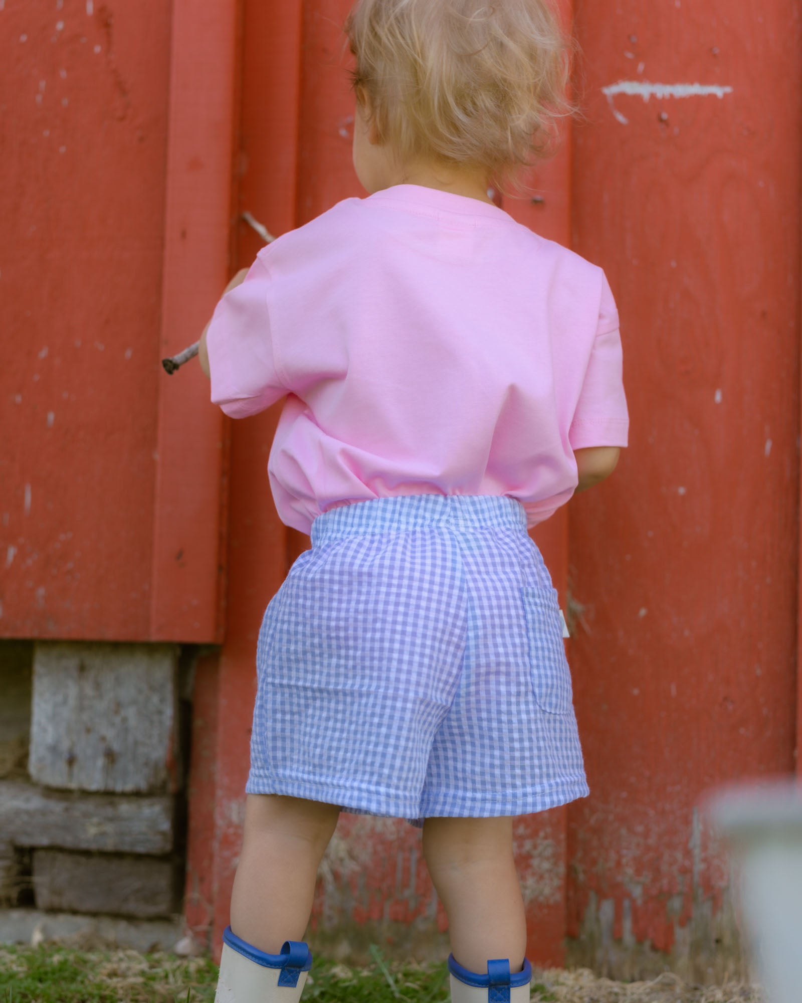 Child wearing a pink shirt and blue checkered shorts standing in front of a red wall.