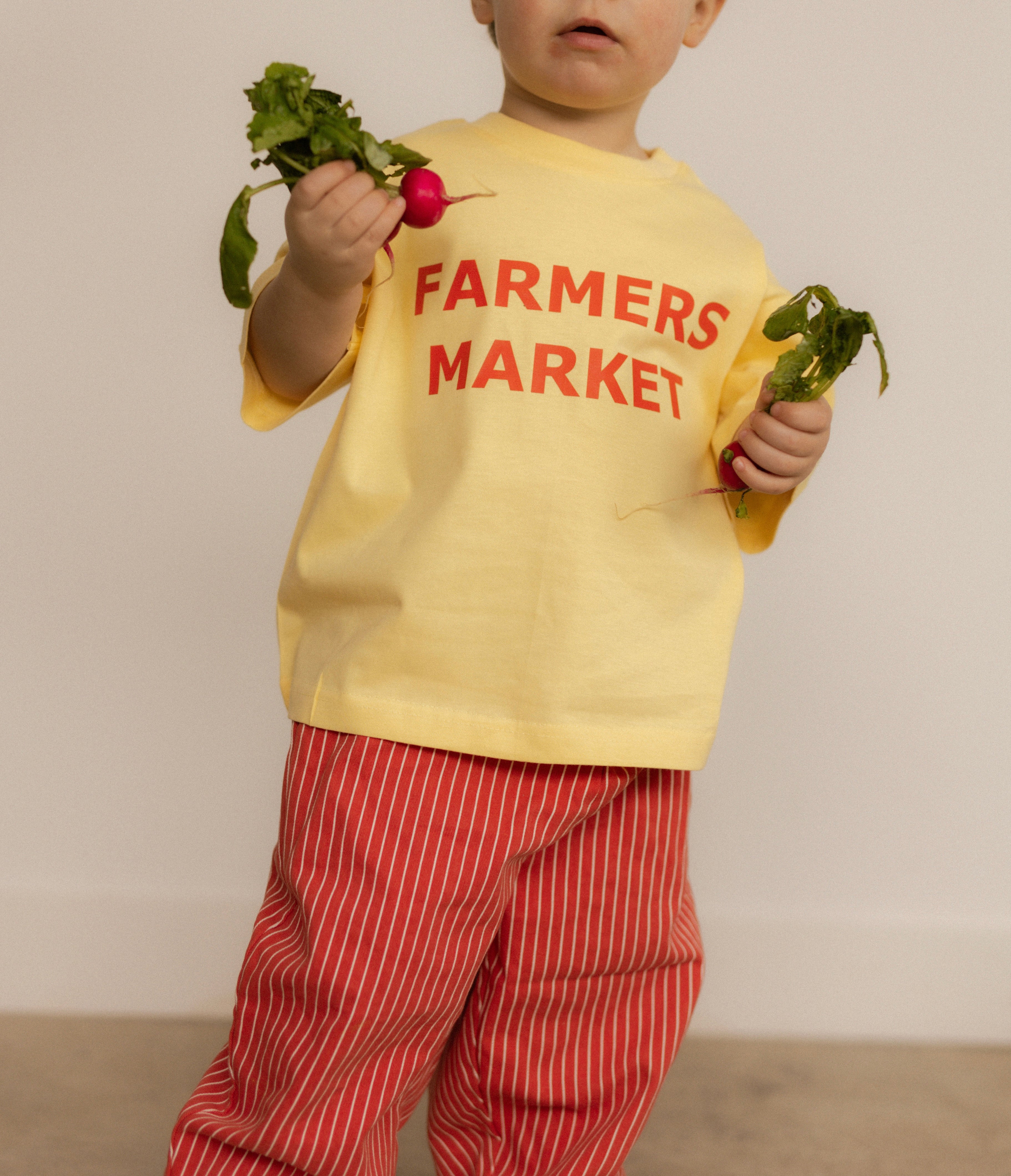 Child wearing a yellow 'Farmers Market' shirt and red stripe pants holding vegetables against a plain background