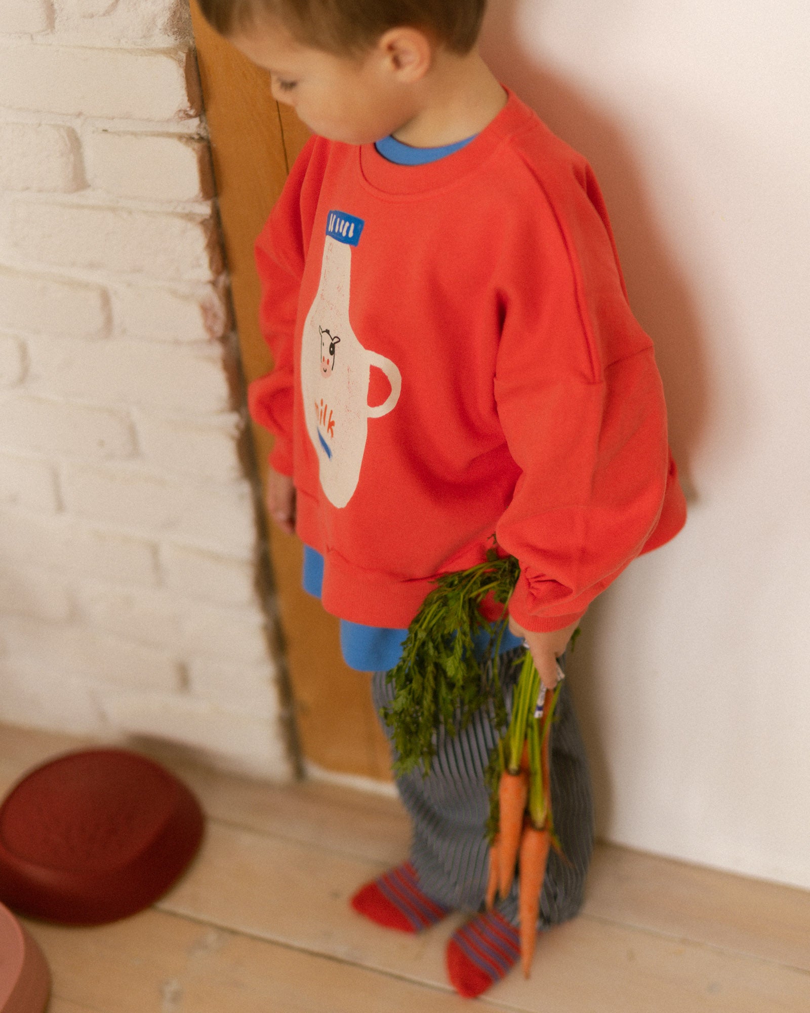 Child in a red sweater with milk bottle on it holding carrots indoors against a white brick wall.