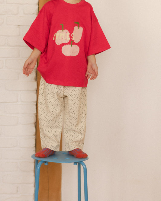 Person wearing a red shirt with graphic design and polka dot pants, standing on a blue stool against a white brick wall.