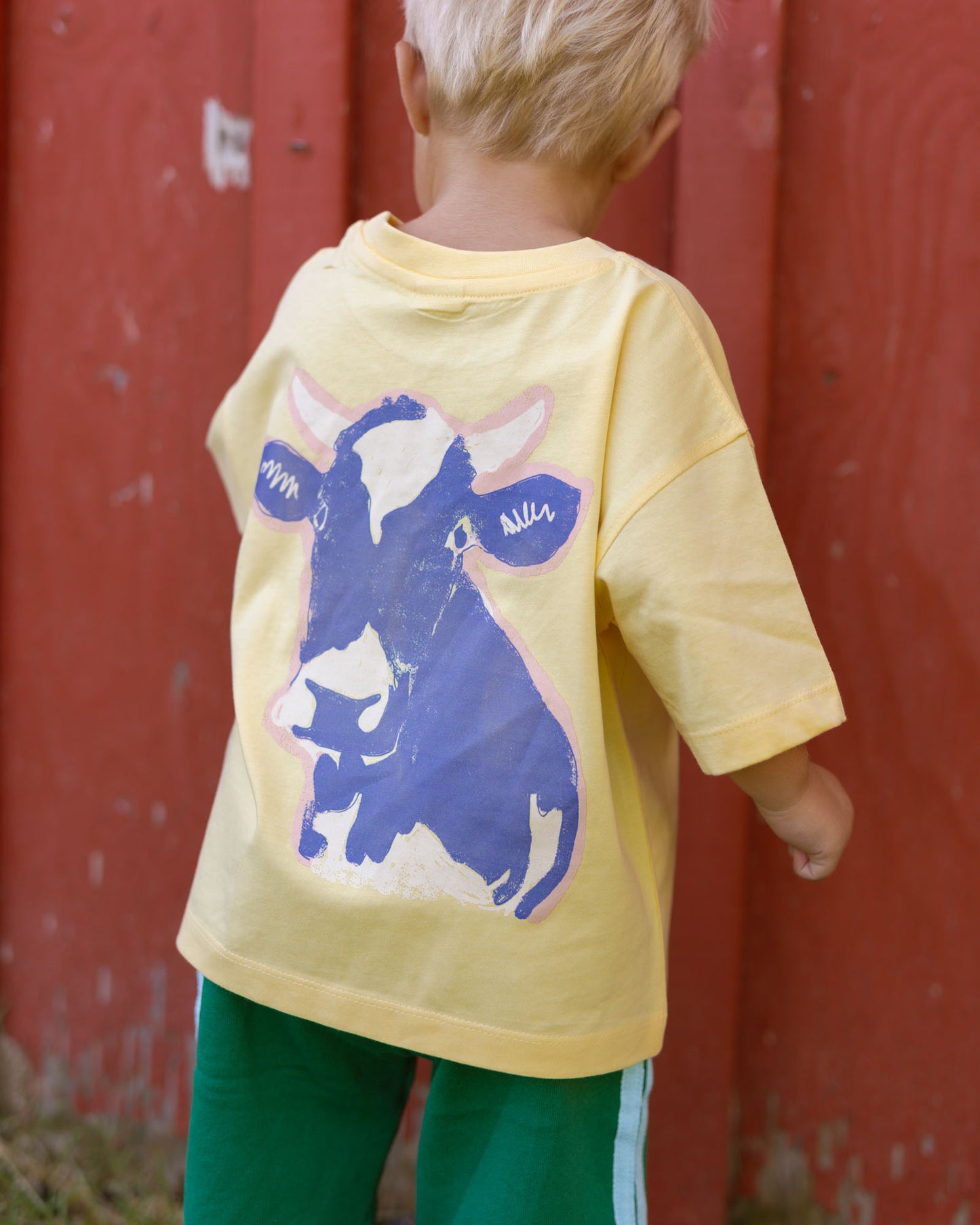 A child viewed from behind wearing a pale yellow oversized T-shirt with a large blue and white cow graphic on the back, paired with green pants, standing in front of a red wooden barn wall.