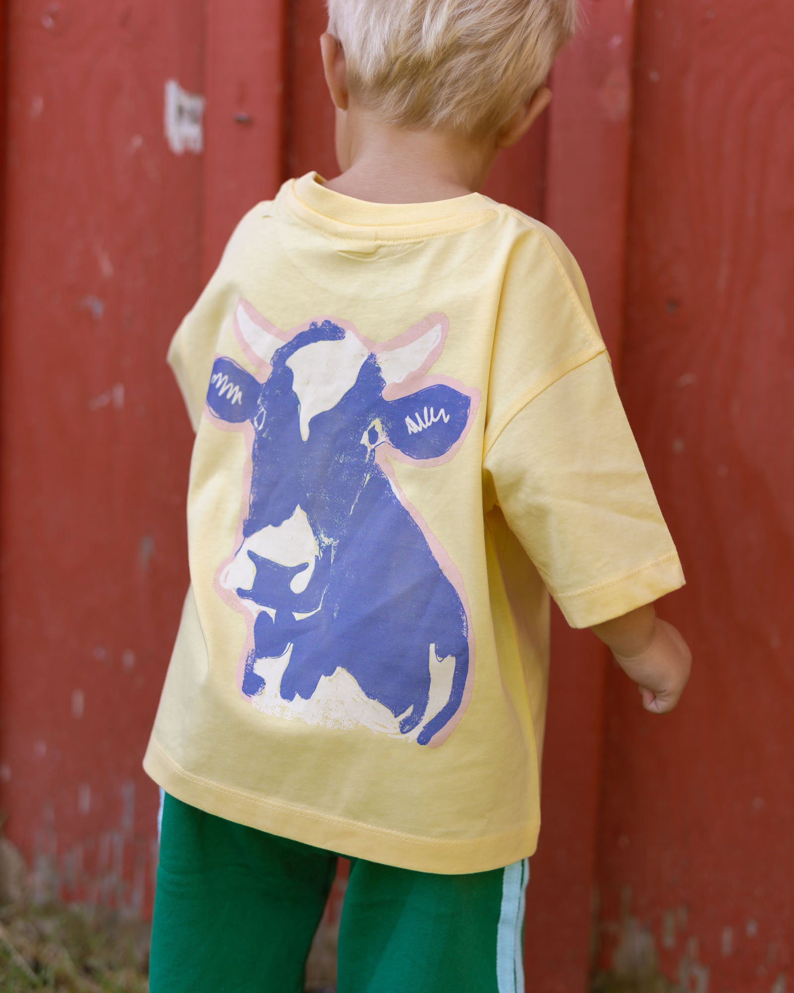 A child viewed from behind wearing a pale yellow oversized T-shirt with a large blue and white cow graphic on the back, paired with green pants, standing in front of a red wooden barn wall.