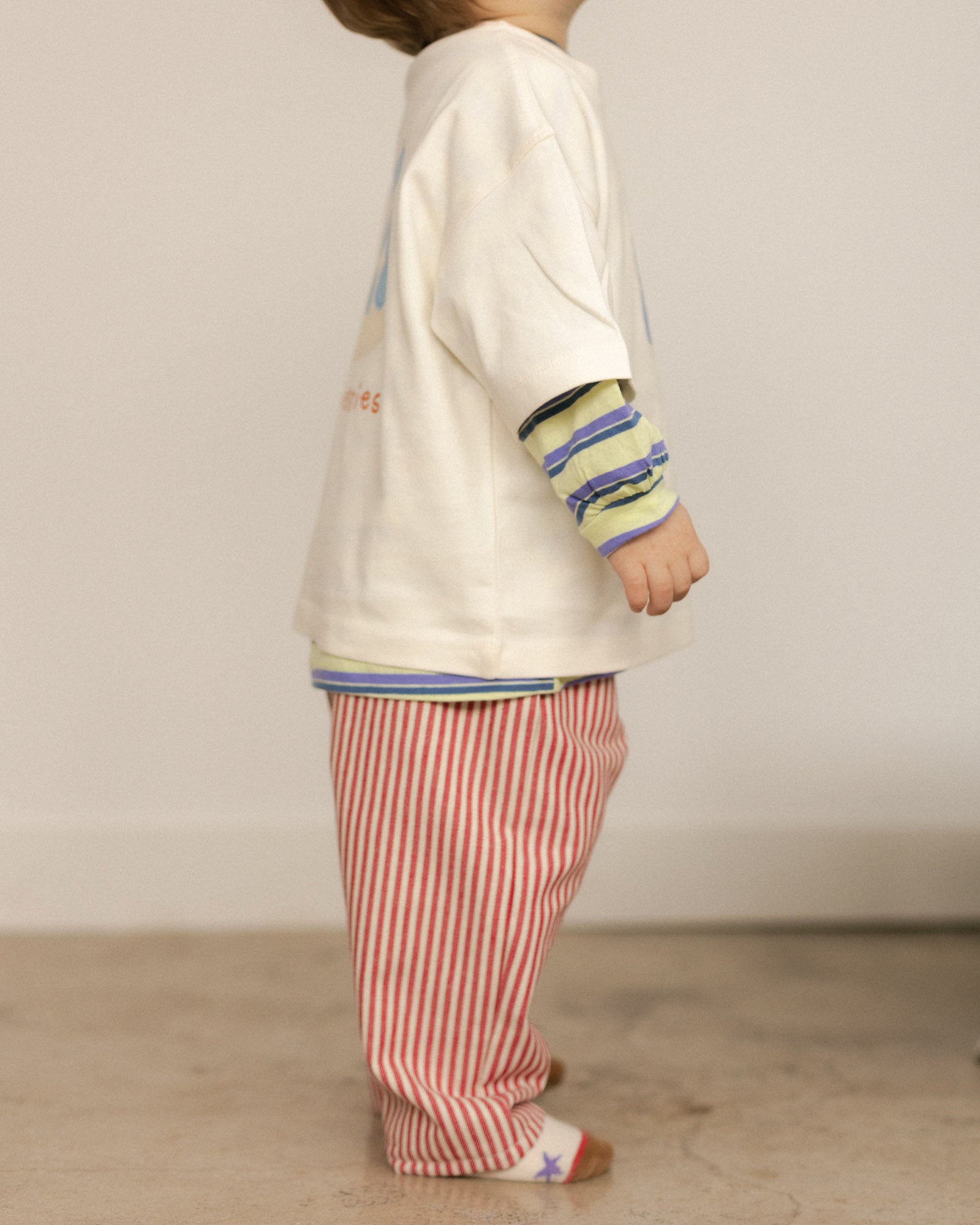 Child wearing a white t-shirt with colorful sleeves and red striped pants against a plain background