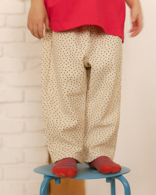 Child wearing a red shirt and beige polka dot pants standing on a blue stool against a white brick wall.