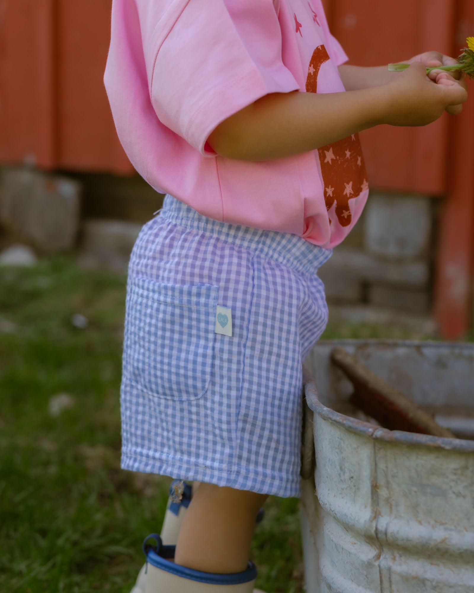 Child wearing a pink shirt and blue checkered shorts standing next to a barrel outdoors.