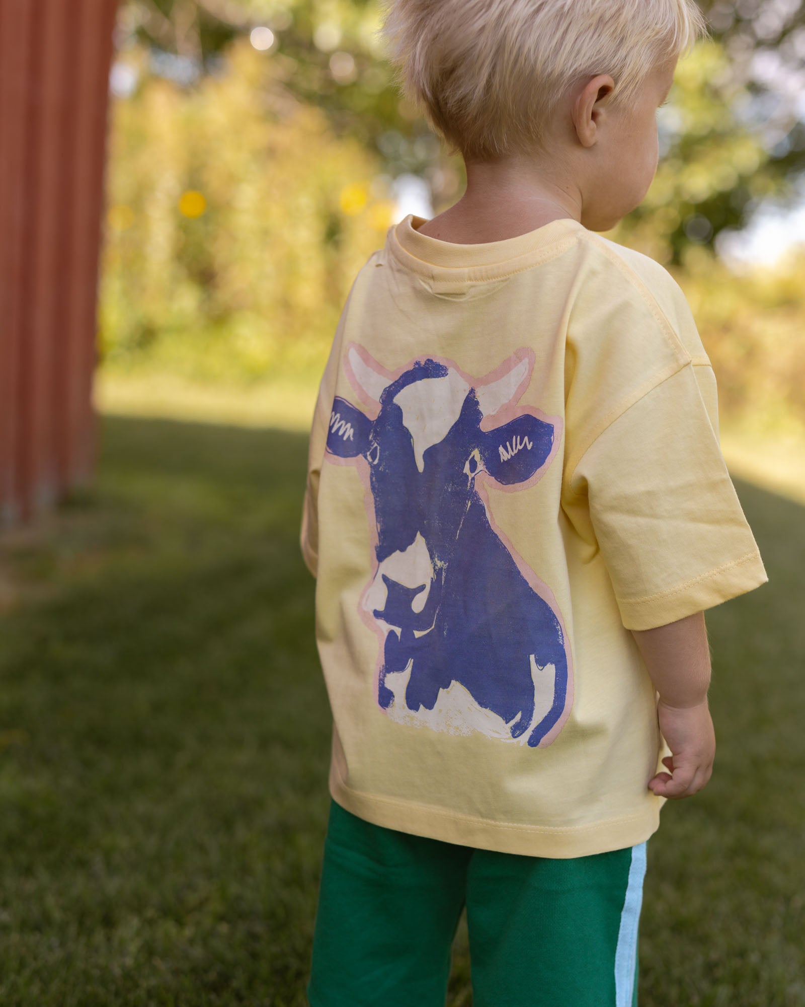 A toddler viewed from behind wearing a pale yellow oversized T-shirt with a large blue and white cow graphic on the back, standing outdoors on grass near a red wooden structure.