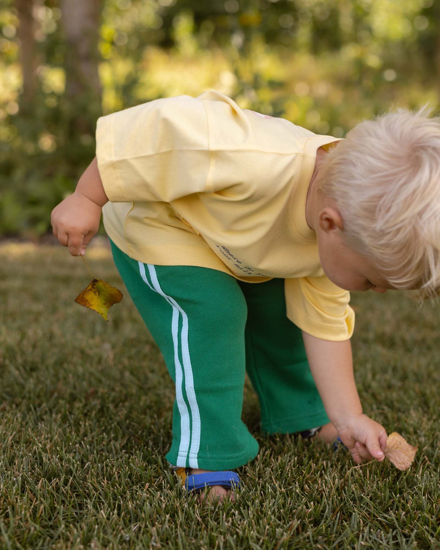 A toddler with blonde hair bends down on a grassy lawn, wearing a pale yellow short-sleeve top and green pants with white side stripes.