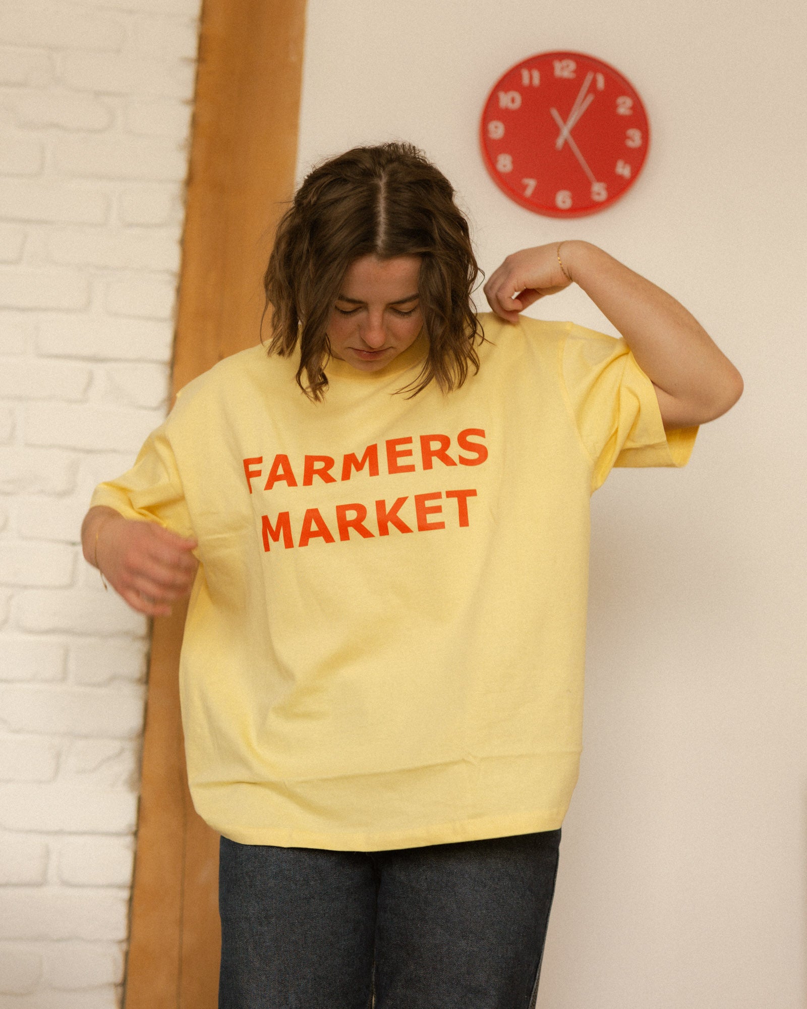 Person wearing a yellow 'Farmers Market' t-shirt in front of a white brick wall with a red clock.