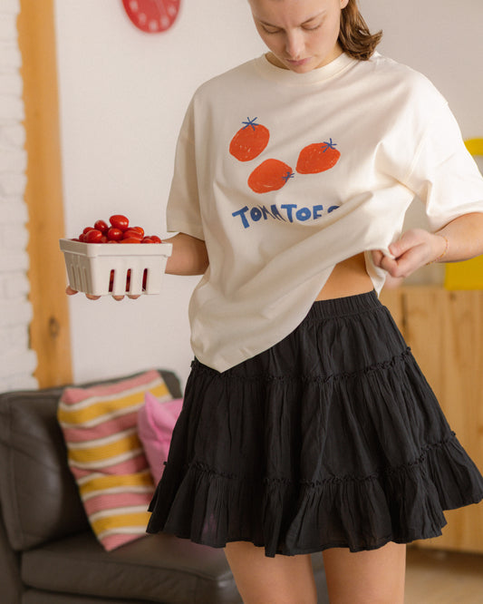 Person wearing a white t-shirt with red tomatoes and text, holding a container of tomatoes in a casual setting.