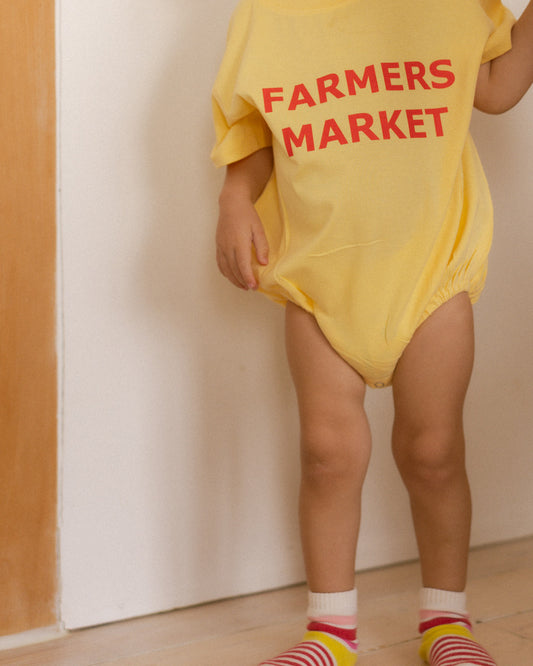 Child wearing a yellow onesie with 'Farmers Market' text against a plain background